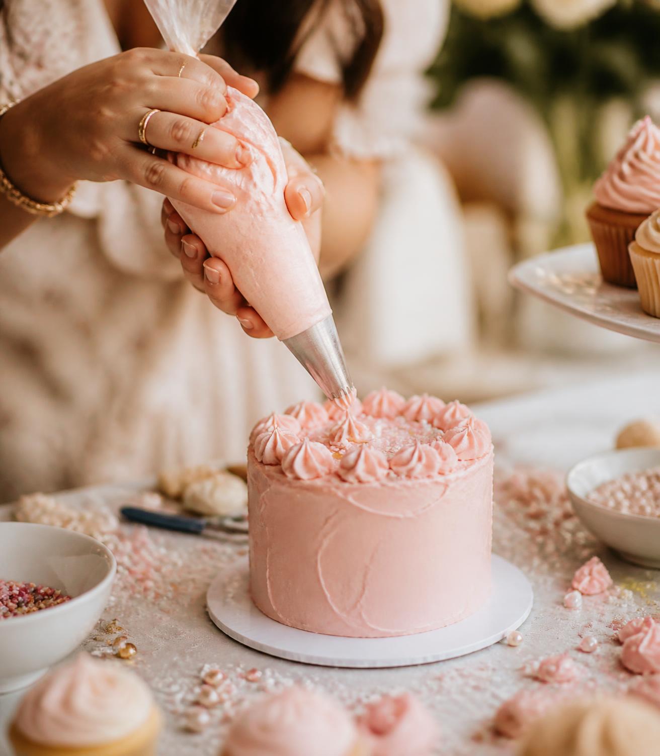 Hands piping soft pink buttercream onto a small cake at a party table